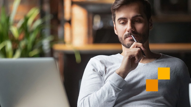 A photo of a young man holding a pen to his mouth as he looks at his laptop.